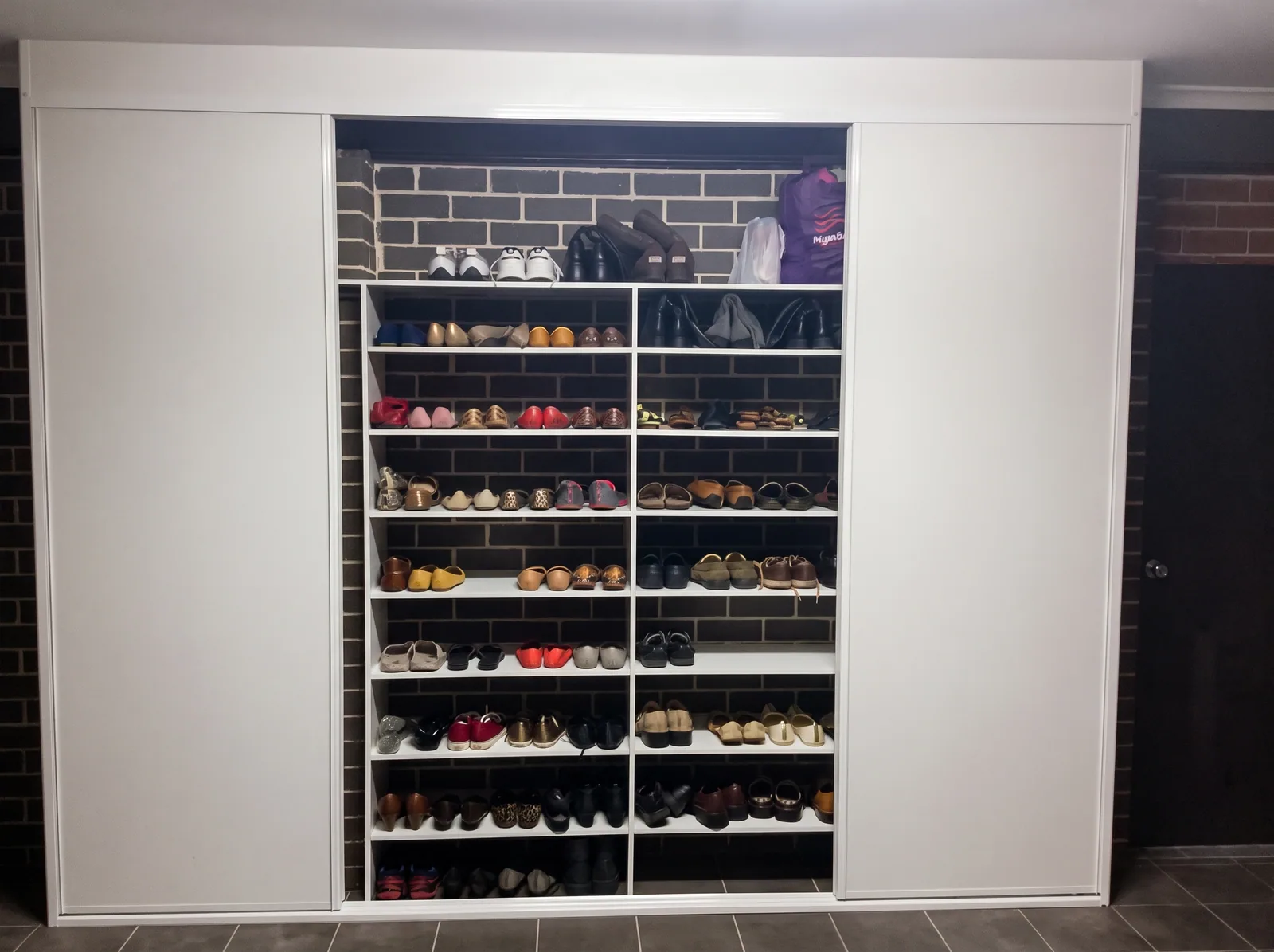 Garage storage wall with white sliding doors either side of floor-to-ceiling shoe shelving against a brick wall, built by Reflections