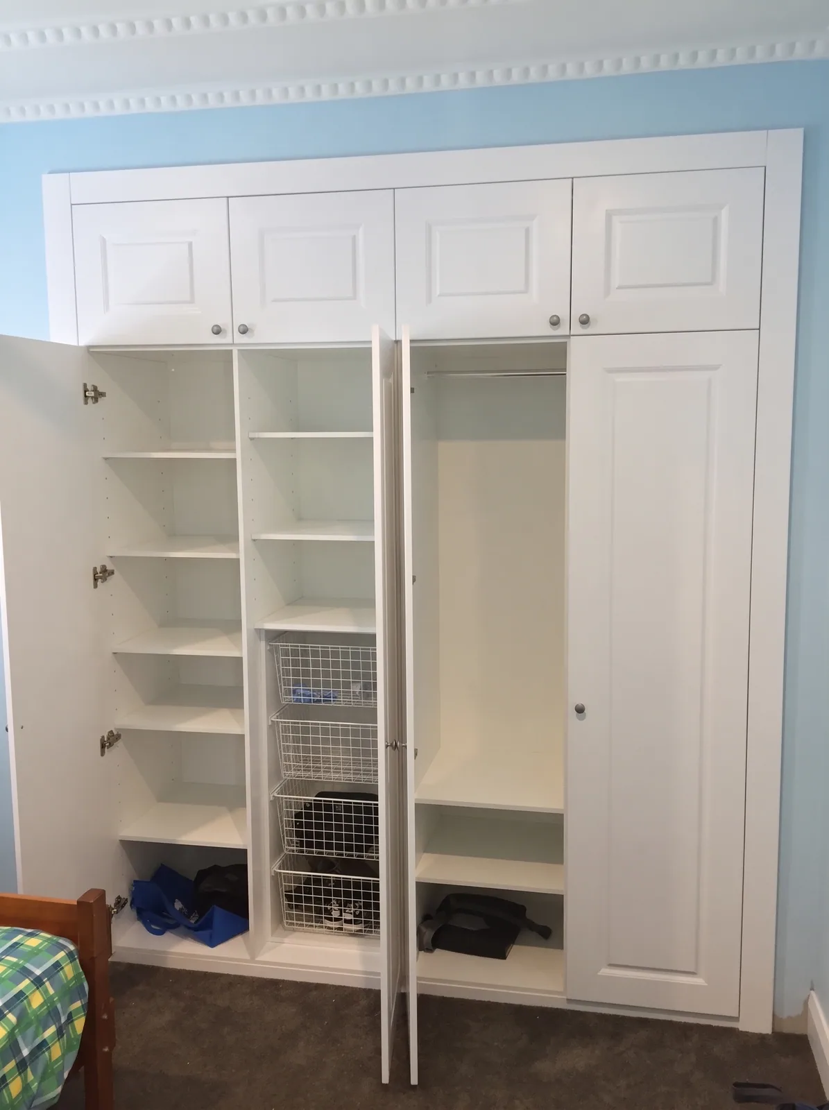White hinged wardrobe with a door open onto shelving and wire baskets, overhead cupboards above, built by Reflections