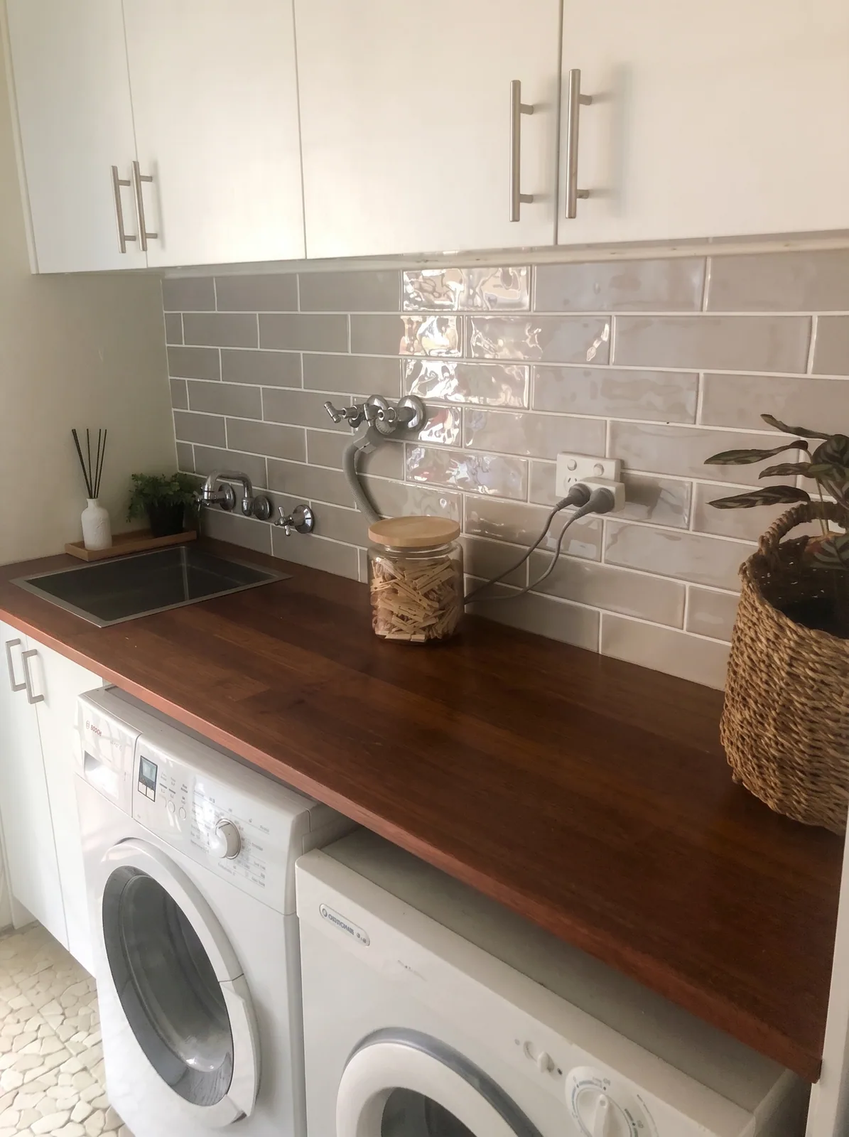 Laundry with a timber-look benchtop and inset sink over two front-loading machines, white overhead cupboards and a tiled splashback, built by Reflections